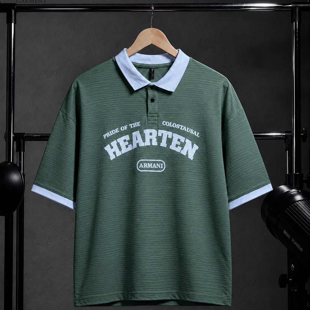 A green short-sleeve polo shirt with a light blue collar and sleeve cuffs, featuring 'HEARTEN' and 'ARMANDO' printed on the front, hanging on a wooden hanger against a dark studio backdrop.