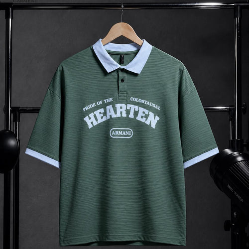 A green short-sleeve polo shirt with a light blue collar and sleeve cuffs, featuring 'HEARTEN' and 'ARMANDO' printed on the front, hanging on a wooden hanger against a dark studio backdrop.