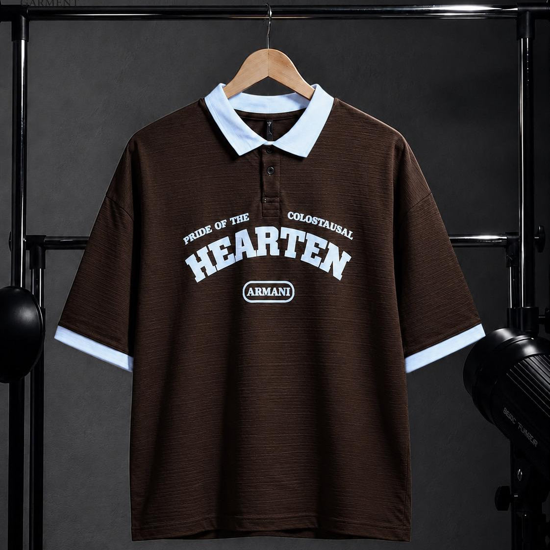 A dark brown short-sleeve polo shirt with a light blue collar and sleeve cuffs, featuring 'HEARTEN' and 'ARMANI' printed in white on the chest, hanging on a wooden hanger against a dark studio backdrop.